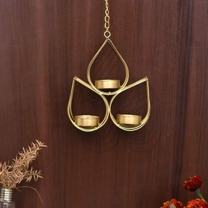 May include: Gold metal candle holder with three teardrop-shaped compartments for tea lights. The holder is suspended from a chain and hangs against a wood background.