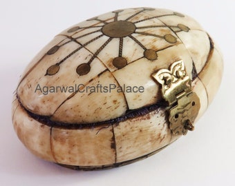 May include: Oval bone trinket box with a brass latch and intricate geometric design. The box is decorated with brass accents and has a light brown color.
