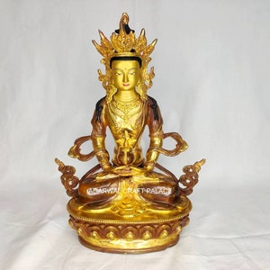 May include: A gold-colored statue of a deity sitting in a meditative pose. The statue is adorned with intricate details and a crown. The deity has a serene expression and is holding a lotus flower in their hands. The statue is sitting on a base with a decorative design.