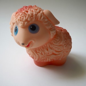 May include: A pink rubber toy lamb with blue eyes and a smiling mouth. The lamb has a fluffy texture and is a classic children's toy.