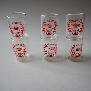 May include: Six clear glass shot glasses stacked in pairs. Each glass has a red logo with the word "КЛИНН" and a stylized image of a building.