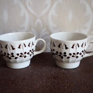 Puede incluir: Dos tazas de té de cerámica blanca con diseños florales marrones y un borde festoneado. Las tazas tienen asas y están sobre una superficie marrón.