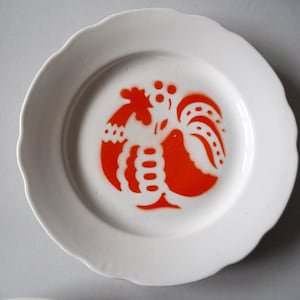 May include: A white ceramic plate with a scalloped edge features a red rooster design in the center.