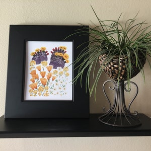 May include: A framed print of a botanical illustration featuring a cluster of orange and yellow California poppies with purple and green foliage. The print is displayed on a black shelf with a green plant in a wicker basket.