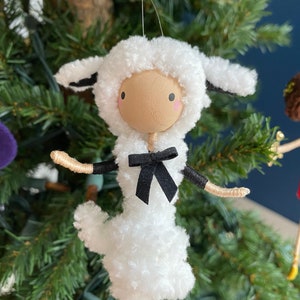 May include: A handmade Christmas ornament shaped like a sheep. The sheep has a white, fluffy body and head, with a light brown face and black eyes. It has a black bow tie and black legs. The ornament is hanging on a Christmas tree.