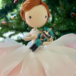 May include: A handmade Christmas ornament featuring a ballerina doll holding a nutcracker. The doll has red hair, a white dress, and a pink fabric flower. The nutcracker is green and gold.