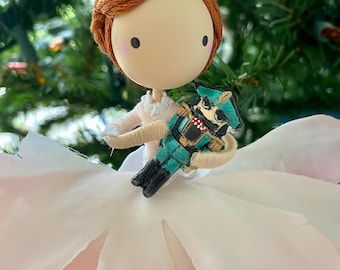 May include: A handmade Christmas ornament featuring a ballerina doll holding a nutcracker. The doll has red hair, a white dress, and a pink fabric flower. The nutcracker is green and gold.