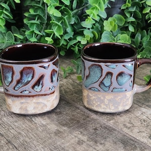 May include: Two ceramic mugs with a unique design. The mugs have a dark brown interior and handle, with a light gray and teal pattern on the upper half. The bottom half of the mugs is a textured beige. The mugs are displayed on a wooden surface.