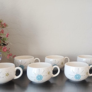 May include: A set of six white ceramic mugs featuring a repeating starburst pattern in shades of blue and green. Each mug has a rounded body and a small handle. The mugs are arranged in a row on a dark surface next to a woven basket of flowers.