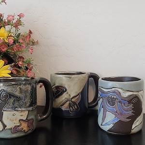 May include: Three ceramic mugs with distinct hand-painted designs. The first mug features a golfer silhouette, the second displays a butterfly motif, and the third shows abstract flowing lines in shades of blue, brown, cream, and dark gray.
