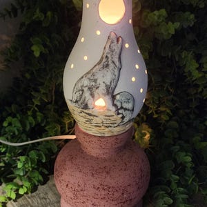 May include: A ceramic lamp with a white, bulbous top section featuring a cutout of a howling wolf and a moon, illuminated from within. The base is a speckled, dusty rose color with a rounded, wide shape. A white power cord extends from the side.
