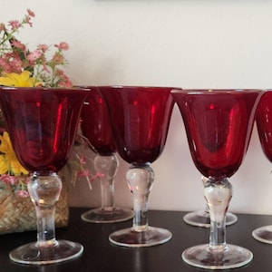 May include: Set of six ruby red wine glasses with clear, decorative stems. The glasses are arranged on a dark surface, with a woven basket of flowers in the background. The glasses are approximately 7 inches tall.