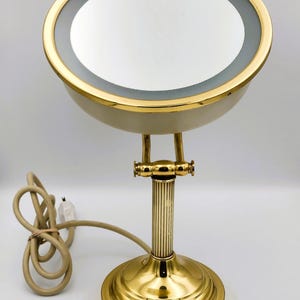 May include: A round, gold-toned vanity mirror with an integrated light ring. The mirror is mounted on a fluted, gold-toned pedestal base with an adjustable hinge. A beige power cord with a small switch extends from the base of the unit.