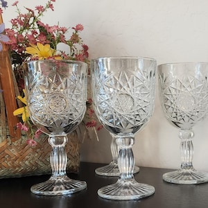 May include: Three clear, patterned wine glasses with starburst designs. The glasses have a stemmed base and are arranged on a dark surface. A woven basket filled with colorful flowers is in the background.