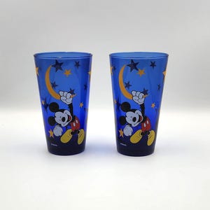 May include: Two blue drinking glasses featuring Mickey Mouse. The glasses have a night sky design with a yellow crescent moon, stars, and Mickey Mouse in a playful pose. The glasses are made of a translucent material.