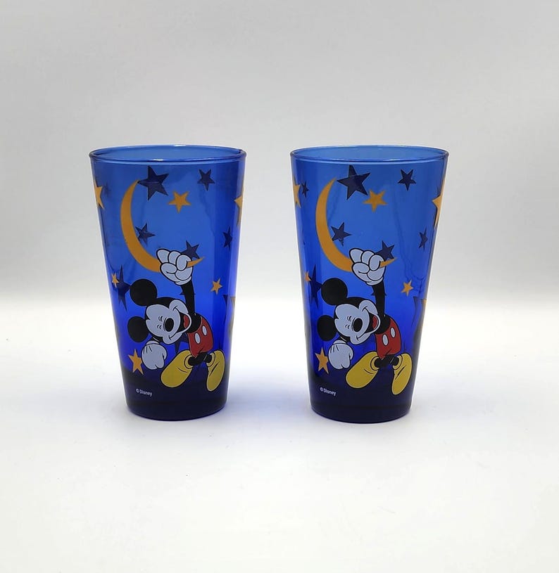 May include: Two blue drinking glasses featuring Mickey Mouse. The glasses have a night sky design with a yellow crescent moon, stars, and Mickey Mouse in a playful pose. The glasses are made of a translucent material.