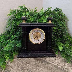May include: A black mantel clock with a rectangular frame and two decorative finials on top. The clock face is circular with a gold-toned background, black Roman numerals, and black hands. The structure is supported by two vertical pillars.