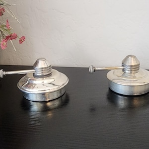 May include: Two silver-colored metal oil cans with a circular base, a domed top, and a thin, straight spout extending from the side. The oil cans have a polished, reflective finish and a tiered, conical cap design at the center of each unit.