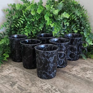 May include: Six black and gray speckled ceramic mugs with unique handle designs. The mugs are arranged on a wooden surface in front of a backdrop of green foliage. The mugs have a marbled appearance.