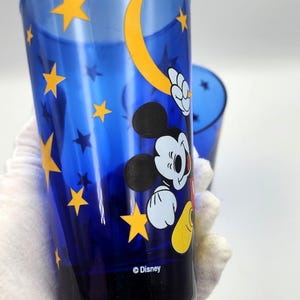 May include: Blue Disney tumbler featuring Mickey Mouse, yellow stars, and a crescent moon design. The tumbler has a cylindrical shape and the Disney logo is visible at the bottom. The glass is held by a gloved hand.