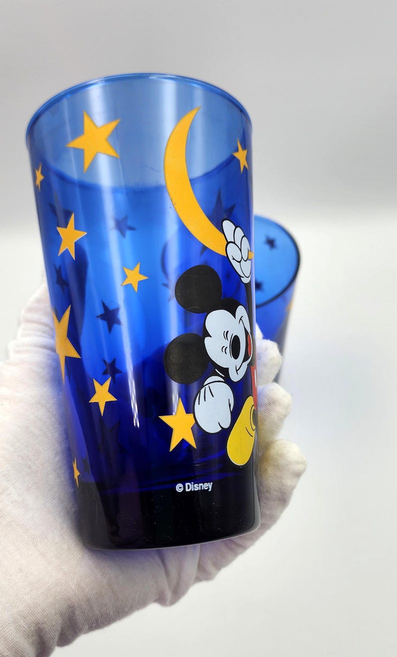 May include: Blue Disney tumbler featuring Mickey Mouse, yellow stars, and a crescent moon design. The tumbler has a cylindrical shape and the Disney logo is visible at the bottom. The glass is held by a gloved hand.