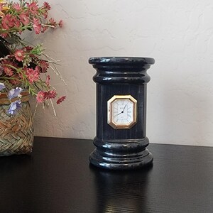 May include: A black cylindrical desk clock with a glossy finish and a column-like shape. The clock face is octagonal with a gold-toned frame, white background, black numerals, and black hands, centered on the front of the black pillar structure.