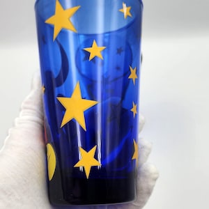May include: A tall, blue glass with yellow star and moon designs. The glass is held by a gloved hand. The glass has a black base and a curved top. The stars are of varying sizes and are evenly spaced around the glass.