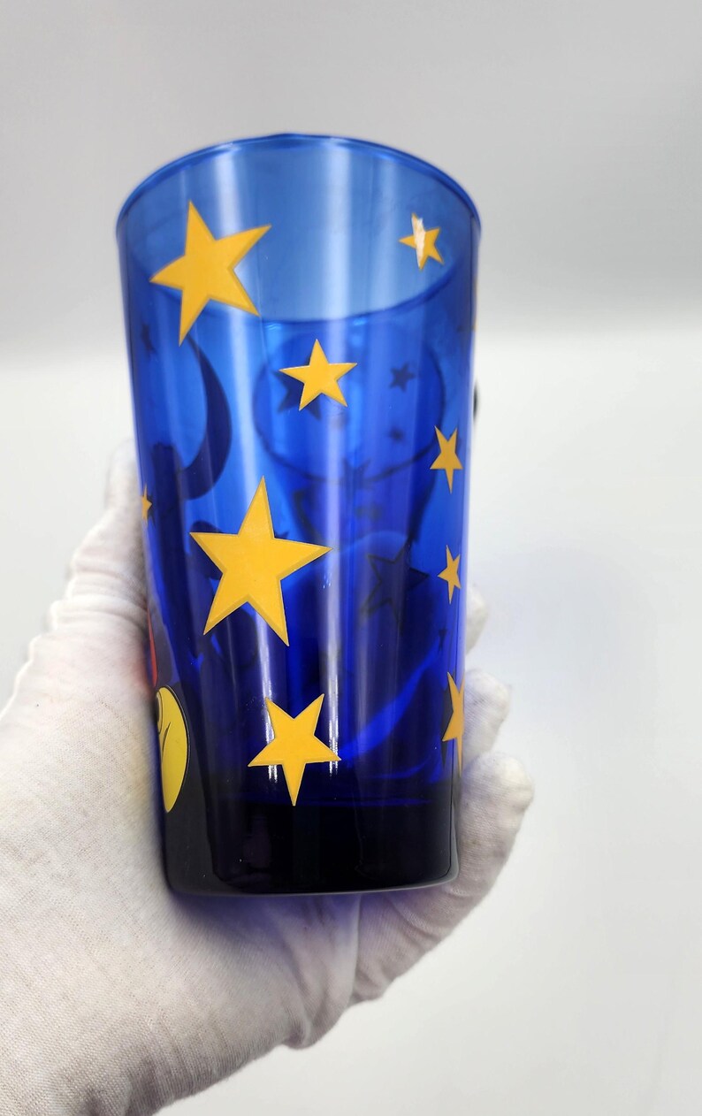 May include: A tall, blue glass with yellow star and moon designs. The glass is held by a gloved hand. The glass has a black base and a curved top. The stars are of varying sizes and are evenly spaced around the glass.