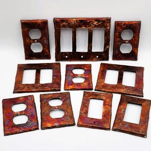 May include: A collection of copper-toned electrical outlet and switch plate covers. The plates have a textured, mottled appearance with shades of red, orange, and purple. Various configurations include single and double outlet covers, and switch plates.