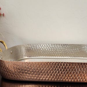 May include: A copper-colored rectangular serving tray with a hammered texture and brass handles. The interior is silver. The tray is on a dark surface, with a basket of flowers in the background. A decorative piece for serving food.