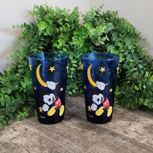 May include: Two blue drinking glasses with a Mickey Mouse design. The glasses feature a dark blue background with yellow stars and a crescent moon. Mickey Mouse is depicted in his classic outfit. The glasses are on a wooden surface.