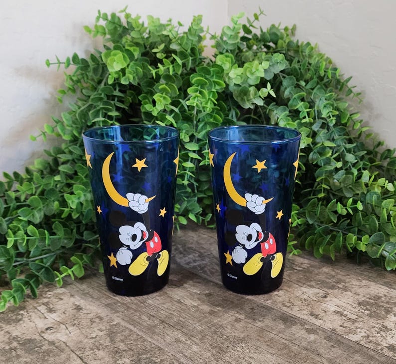 May include: Two blue drinking glasses with a Mickey Mouse design. The glasses feature a dark blue background with yellow stars and a crescent moon. Mickey Mouse is depicted in his classic outfit. The glasses are on a wooden surface.