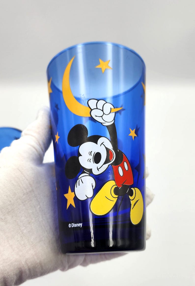 May include: Blue drinking glass featuring Mickey Mouse with a yellow crescent moon and stars. Mickey is wearing red shorts, yellow shoes, and white gloves. The Disney logo is at the bottom.