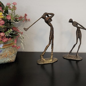 May include: Two bronze golf figurines, one in a follow-through pose and the other in a backswing. A basket of flowers with yellow and pink blooms sits to the left. The figurines are on a black surface.