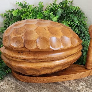 May include: A handcrafted wooden nutcracker set, featuring a large, intricately carved bowl with a lid, a base, and a small mallet. The wood has a warm, natural tone, and the design is reminiscent of a clam shell. The set is displayed against a backdrop of green foliage.
