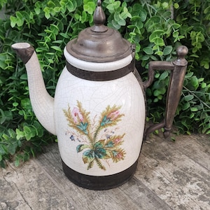 May include: A white ceramic teapot with a floral design and a silver lid. The teapot has a crackled glaze and a black metal band around the top and bottom.