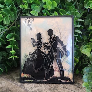 May include: A framed silhouette art piece featuring a couple in vintage attire. The black silhouettes are set against a soft, pastel-colored background. The frame is black and rectangular, with a classic, romantic theme.