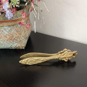 May include: Ornate brass sugar tongs with a floral design. The tongs are open, displaying the tweezer-like ends. The handle features detailed leaf and flower patterns. The item is on a black surface.