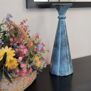 May include: A tall, conical candle holder with a textured, weathered blue finish. The object features a flared top rim and a tapered body with vertical ridges. Beside it sits a woven basket filled with a variety of pink, yellow, and blue flowers.