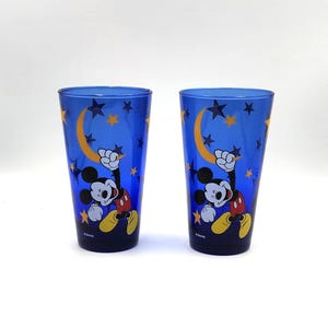 May include: Two blue drinking glasses featuring Mickey Mouse with a crescent moon and star design. The glasses have a tall, cylindrical shape and are decorated with a cartoon image of Mickey Mouse. The background is a dark blue color.