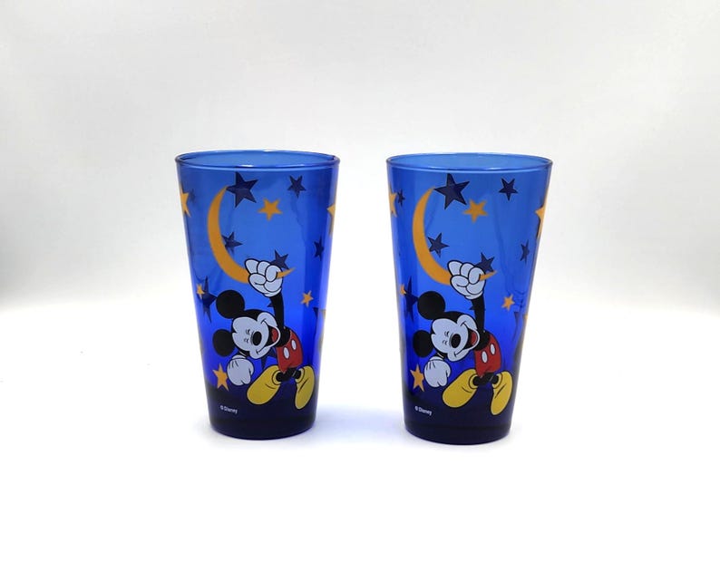 May include: Two blue drinking glasses featuring Mickey Mouse with a crescent moon and star design. The glasses have a tall, cylindrical shape and are decorated with a cartoon image of Mickey Mouse. The background is a dark blue color.