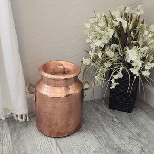 May include: A white ceramic mug with a black handle and the text "But First, Coffee" in a bold, black font. The mug is set against a blurred background, suggesting a cozy setting.