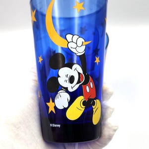 May include: A blue plastic tumbler featuring Mickey Mouse with a yellow crescent moon and stars design. The Disney-branded cup has a black base and a handle. The image shows Mickey Mouse in a dynamic pose.