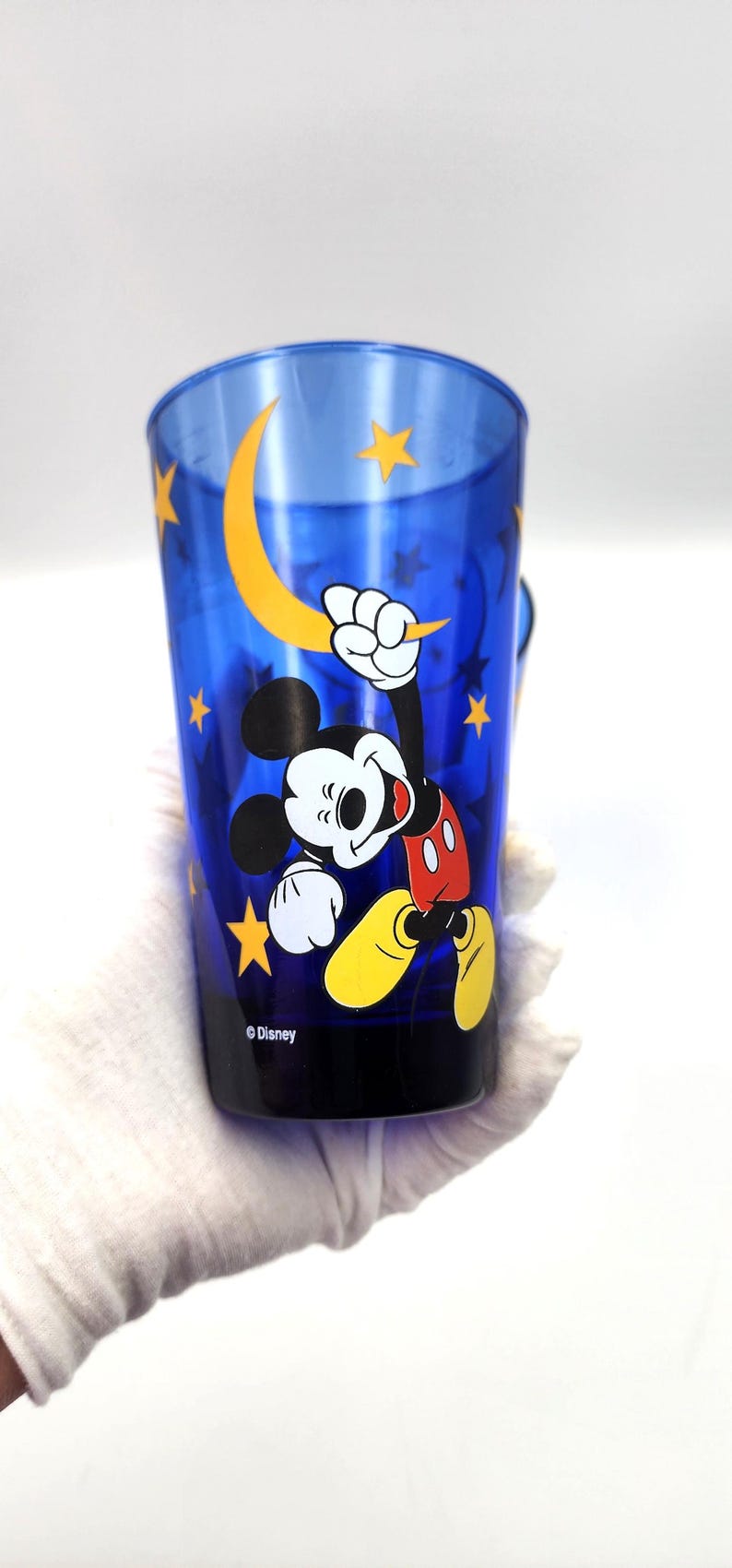 May include: A blue plastic tumbler featuring Mickey Mouse with a yellow crescent moon and stars design. The Disney-branded cup has a black base and a handle. The image shows Mickey Mouse in a dynamic pose.