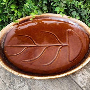 May include: Oval-shaped brown ceramic serving dish with a leaf design. The dish has a glossy finish and a textured, light brown rim. The background features green foliage and a wooden surface.