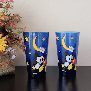 May include: Two blue drinking glasses featuring Mickey Mouse holding a crescent moon. The glasses have a night sky design with yellow stars. The Disney logo is at the bottom of each glass.