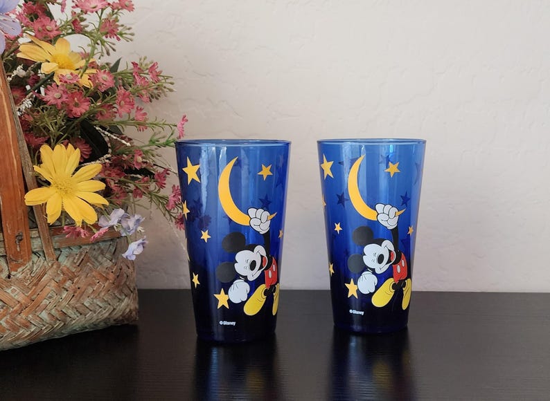 May include: Two blue drinking glasses featuring Mickey Mouse holding a crescent moon. The glasses have a night sky design with yellow stars. The Disney logo is at the bottom of each glass.