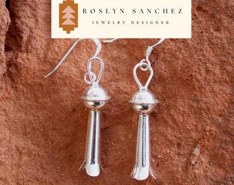 May include: A pair of silver dangle earrings with a teardrop shape and a rounded top. The earrings are on a red-brown textured surface.