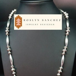 May include: A silver necklace with alternating round and oval beads. The necklace is displayed on a black mannequin bust. The label on the bust reads "Roslyn Sanchez Jewelry Designer".