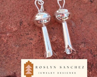 May include: A pair of silver earrings with a floral design. Each earring features a round top and a long, bell-shaped bottom. The earrings are displayed on a reddish-brown surface. The text "ROSLYN SANCHEZ JEWELRY DESIGNER" is at the bottom.